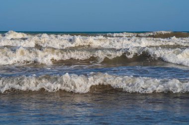 Denizde hafif bir fırtına. Beyaz köpüklü ve beyaz kapaklı dalgalar kumlu bir plajın kıyısında yuvarlanıyor. İspanya 'da tatil köyü. Deniz kıyısının güzel manzarası