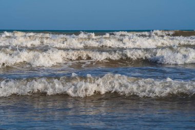 Denizde hafif bir fırtına. Beyaz köpüklü ve beyaz kapaklı dalgalar kumlu bir plajın kıyısında yuvarlanıyor. İspanya 'da tatil köyü. Deniz kıyısının güzel manzarası