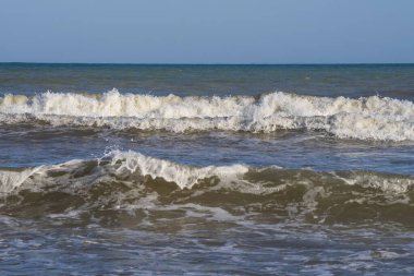 Denizde hafif bir fırtına. Beyaz köpüklü ve beyaz kapaklı dalgalar kumlu bir plajın kıyısında yuvarlanıyor. İspanya 'da tatil köyü. Deniz kıyısının güzel manzarası