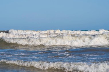 Denizde hafif bir fırtına. Beyaz köpüklü ve beyaz kapaklı dalgalar kumlu bir plajın kıyısında yuvarlanıyor. İspanya 'da tatil köyü. Deniz kıyısının güzel manzarası