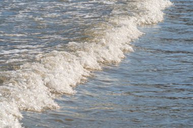 Güzel, köpüklü ve beyaz kapaklı tek bir dalga kıyıya vuruyor. Deniz suyu güneşte parlıyor. Sahildeki deniz dalgası