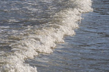 Güzel, köpüklü ve beyaz kapaklı tek bir dalga kıyıya vuruyor. Deniz suyu güneşte parlıyor. Sahildeki deniz dalgası