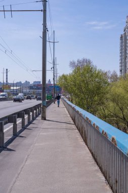 İlkbaharda yoğun bir yol boyunca nehrin üzerindeki köprünün kaldırımında. Modern mimari. Şehrin köprüsü.