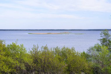 Mavi gökyüzünün altında sazlıklar ve yeşil ağaçlarla sakin bir nehrin manzarası.