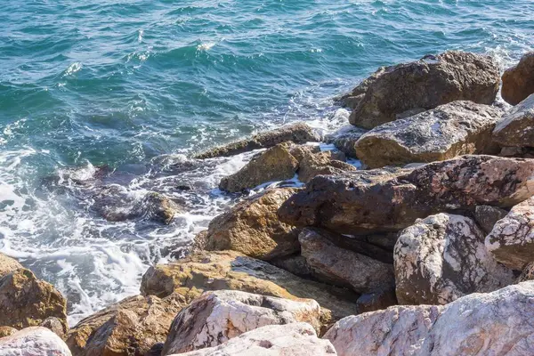 Dalgalar halinde akan turkuaz deniz suyuyla kayalık kıyılar ve kalın beyaz deniz köpüğü oluşturur..