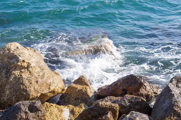 Dalgalar halinde akan turkuaz deniz suyuyla kayalık kıyılar ve kalın beyaz deniz köpüğü oluşturur..