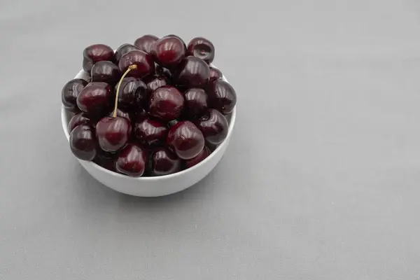 Taze yaz meyvelerinin yakın plan gri arka planında beyaz bir kasede olgun siyah kirazlar.