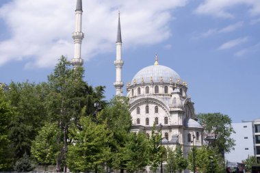 İstanbul 'daki Dolmabahe Camii. Yeşil ağaçların arkasında zarif kubbeleri ve minareleri olan tarihi Osmanlı camisi. Açık mavi gökyüzü ve güneş ışığı mimari güzelliği vurguluyor.