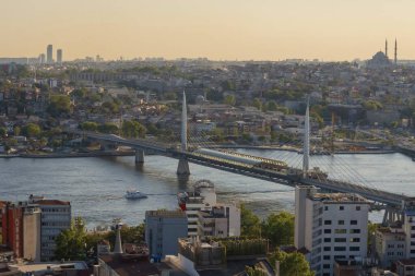 İstanbul 'daki Haliç Köprüsü' nü feribot, eski binalar ve arka planda tarihi Valens Su kemeri ile geçen modern Hali Metro Köprüsü. 20 Mayıs 2025 'te gün batımında yakalandılar..