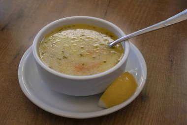 Yanında otlar, havuçlar ve limon dilimleri olan seramik bir kasede taze hazırlanmış Türk çorbası. Doğal ışık altında ahşap bir masada yakalanmış. Yemek ve restoran teması için ideal..