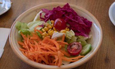 İçinde öğütülmüş havuçlar, kiraz domatesleri, mısır çekirdekleri, salatalık dilimleri, buzdağı marulu ve ahşap bir yüzey üzerinde doğranmış kırmızı lahana bulunan seramik bir kasede taze sebze salatasının yakın çekimi..