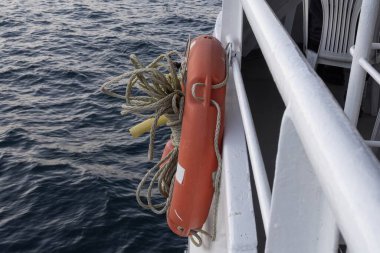 Bir geminin korkuluklarından sarkan ipli turuncu can simidi, açık denizin arka planına kurulmuş. Deniz güvenliği, seyahat, denizcilik ve acil kurtarma konseptleri için ideal..