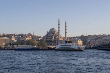 İstanbul gün batımında Yeni Cami, Galata Köprüsü ve boğazdan geçen feribotlarla kıyı manzarası. Osmanlı mimarisi, canlı pazarlar ve deniz faaliyetlerini harmanlayan pitoresk şehir manzarası. 19 Mayıs 2025