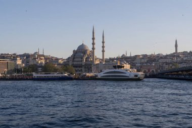 İstanbul gün batımında Yeni Cami, Galata Köprüsü ve boğazdan geçen feribotlarla kıyı manzarası. Osmanlı mimarisi, canlı pazarlar ve deniz faaliyetlerini harmanlayan pitoresk şehir manzarası. 19 Mayıs 2025