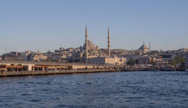 İstanbul gün batımında Yeni Cami, Galata Köprüsü ve boğazdan geçen feribotlarla kıyı manzarası. Osmanlı mimarisi, canlı pazarlar ve deniz faaliyetlerini harmanlayan pitoresk şehir manzarası. 19 Mayıs 2025