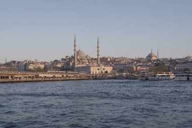 İstanbul gün batımında Yeni Cami, Galata Köprüsü ve boğazdan geçen feribotlarla kıyı manzarası. Osmanlı mimarisi, canlı pazarlar ve deniz faaliyetlerini harmanlayan pitoresk şehir manzarası. 19 Mayıs 2025