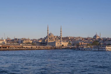 İstanbul gün batımında Yeni Cami, Galata Köprüsü ve boğazdan geçen feribotlarla kıyı manzarası. Osmanlı mimarisi, canlı pazarlar ve deniz faaliyetlerini harmanlayan pitoresk şehir manzarası. 19 Mayıs 2025