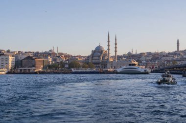İstanbul gün batımında Yeni Cami, Galata Köprüsü ve boğazdan geçen feribotlarla kıyı manzarası. Osmanlı mimarisi, canlı pazarlar ve deniz faaliyetlerini harmanlayan pitoresk şehir manzarası. 19 Mayıs 2025