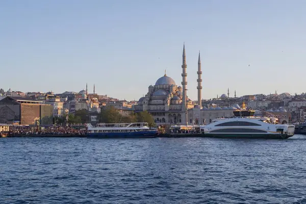 İstanbul gün batımında Yeni Cami, Galata Köprüsü ve boğazdan geçen feribotlarla kıyı manzarası. Osmanlı mimarisi, canlı pazarlar ve deniz faaliyetlerini harmanlayan pitoresk şehir manzarası. 19 Mayıs 2025