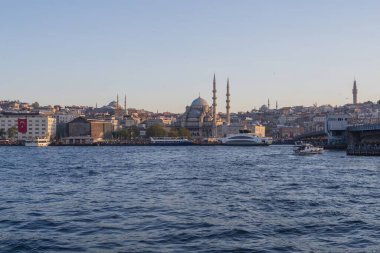 İstanbul gün batımında Yeni Cami, Galata Köprüsü ve boğazdan geçen feribotlarla kıyı manzarası. Osmanlı mimarisi, canlı pazarlar ve deniz faaliyetlerini harmanlayan pitoresk şehir manzarası. 19 Mayıs 2025