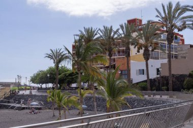 Güneşli bir Tenerife plajında volkanik kayalar, siyah kum ve şemsiyelerle huzurlu deniz manzarası. Palmiye ağaçlarının altında dinlenen ve Atlantik Okyanusu kıyısında sıcak havanın tadını çıkaran turistler..