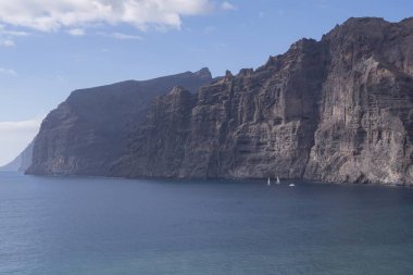 Okyanus dalgaları Tenerife 'deki Los Gigantes kayalıklarının altında volkanik plajı yıkıyor. Atlantik suyuyla güneşli deniz manzarası, sakin ufuk ve dramatik kayalar. Seyahat ve doğa tasarım temaları için ideal.