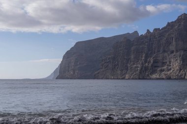 Okyanus dalgaları Tenerife 'deki Los Gigantes kayalıklarının altında volkanik plajı yıkıyor. Atlantik suyuyla güneşli deniz manzarası, sakin ufuk ve dramatik kayalar. Seyahat ve doğa tasarım temaları için ideal.