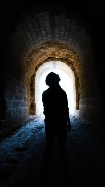 Gizemli bir siluet, loş bir taş tünelde, girişte parlayan ışıkla dikilirken yukarı bakıyor..