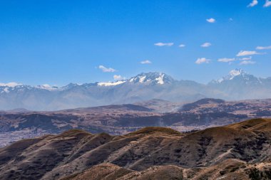 Peru 'daki And dağlarının manzarası, ön planda kuru otlaklar, orta alanda engebeli zirveler ve parlak mavi gökyüzünün altında karla kaplı dağlar.