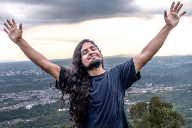 Man with long hair and a beard raising his arms in joy while standing on a mountain top with a scenic valley in the background. A moment of peace and freedom. clipart