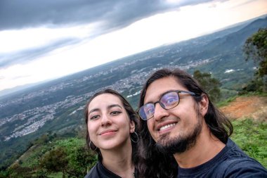 Gün batımının tadını çıkaran gülümseyen bir çift, sıcak ışıklandırmayla bir dağ ortamında selfie çekiyorlar. Bir anlık mutluluk ve bağlantı.