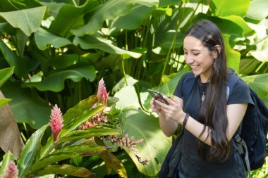 A young woman takes a photo of a pink tropical flower using her smartphone, surrounded by lush green foliage clipart