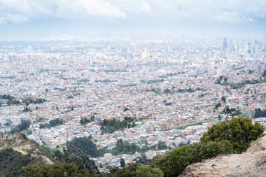 Bogota 'nın panoramik hava manzarası, karışık alçak katlı ve yüksek katlı binalar, yeşil alanlar ve bulutlu bir gökyüzünün altında uzak bir ufuk çizgisi.