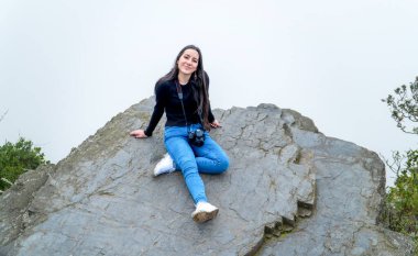 Gülümseyen genç bir kadın arka planda sisli yeşil bir vadiyle kayalık bir uçurumda oturuyor. Doğada barışçıl açık hava macerası