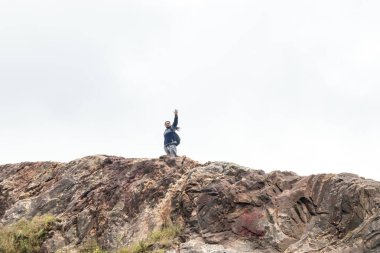 Bir adam engebeli bir kaya oluşumunun üzerinde duruyor, kameraya el sallıyor. Bulutlu gökyüzü dağın toprak tonlarıyla çelişiyor.