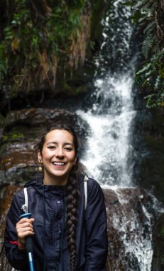 Doğanın ortasında şelalenin yanında yürüyüş yapan bir kadın. Genç bayan gezgin - şelalenin yanındaki turist. Şelalenin yanında yürüyüş direkleri olan bir kadın.