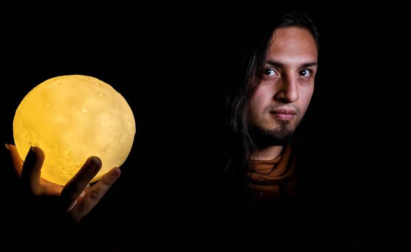 Portrait of a man in dramatic lighting holding an illuminated moon lamp, creating a mystical and surreal atmosphere against a black background