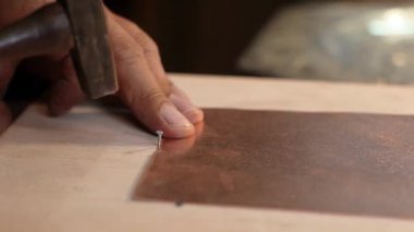 Bir zanaatkar, ahşap bir yüzeyde bakır bir çiviye küçük bir çivi çakmak için çekiç kullanır. Metal işleme hassasiyetini gösterir..