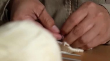 Beyaz yün iplik ve ahşap masada metal iğnelerle örgü ören ellerin yakın çekimi..