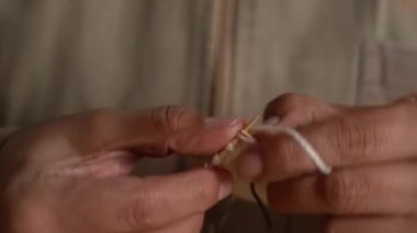 Beyaz yün iplik ve ahşap masada metal iğnelerle örgü ören ellerin yakın çekimi..
