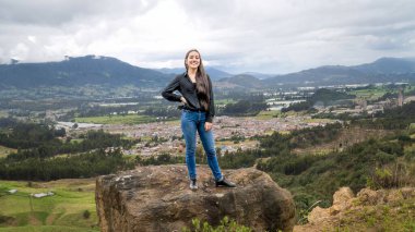 Genç bir kadın, arka planda panoramik bir vadi, kasaba ve dağ manzaralı bir kayanın üzerinde güven içinde poz veriyor. Bulutlu gökyüzü manzaraya derinlik katar.