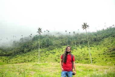 Kolombiya 'nın ikonik gökdelenleri ve sisli yeşil tepelerle çevrili gür Cocora Vadisi' nde kırmızı gömlekli gülümseyen bir gezgin duruyor.