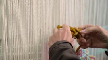 Renkli iplikleri dokuma tezgahına bağlayan ellerin yakın çekimi, geleneksel tekstil yaratılışının karmaşık sürecini vurguluyor.