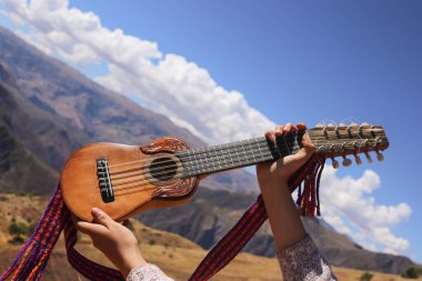 Geleneksel bir Andean charango bir kişi tarafından manzaralı bir dağ zeminine karşı soyulur. Enstrümanda karmaşık oymalar ve renkli dokuma kayışlar yer alıyor.