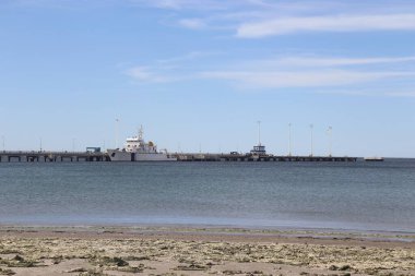 Durgun sulara uzanan bir rıhtım manzarası modern sokak lambalarıyla çevrelenmiş, parlak mavi gökyüzü altında, limanlı bir gemiyle.