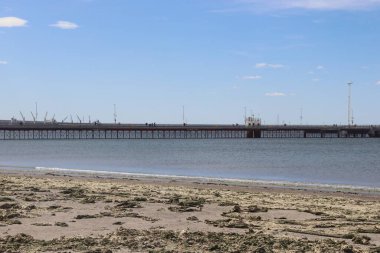 Durgun sulara uzanan bir rıhtım manzarası modern sokak lambalarıyla çevrelenmiş, parlak mavi gökyüzü altında, limanlı bir gemiyle.