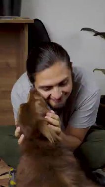 Neşeli bir adam evde otururken kahverengi bir köpek sevgiyle yüzünü yalıyor, karşılıklı şefkat gösteriyor..