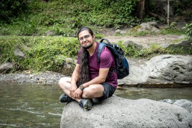 Latin adam dağa gidiyor. Erkek yürüyüşçü doğada oturuyor. İnsan nehirde dinlenir ve eğlenir.