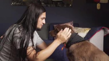 Uzun saçlı bir adam, sıcak, samimi bir oturma odasında sıcak ve samimi bir anı paylaşan kahverengi köpeğin yüzünü sevgiyle tutuyor..