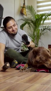 Evcil hayvan yatağında dinlenen kahverengi bir köpeği tımar etmek için saç kurutma makinesi kullanan bir adam sakin ve sevgi dolu bir anı paylaşıyor..